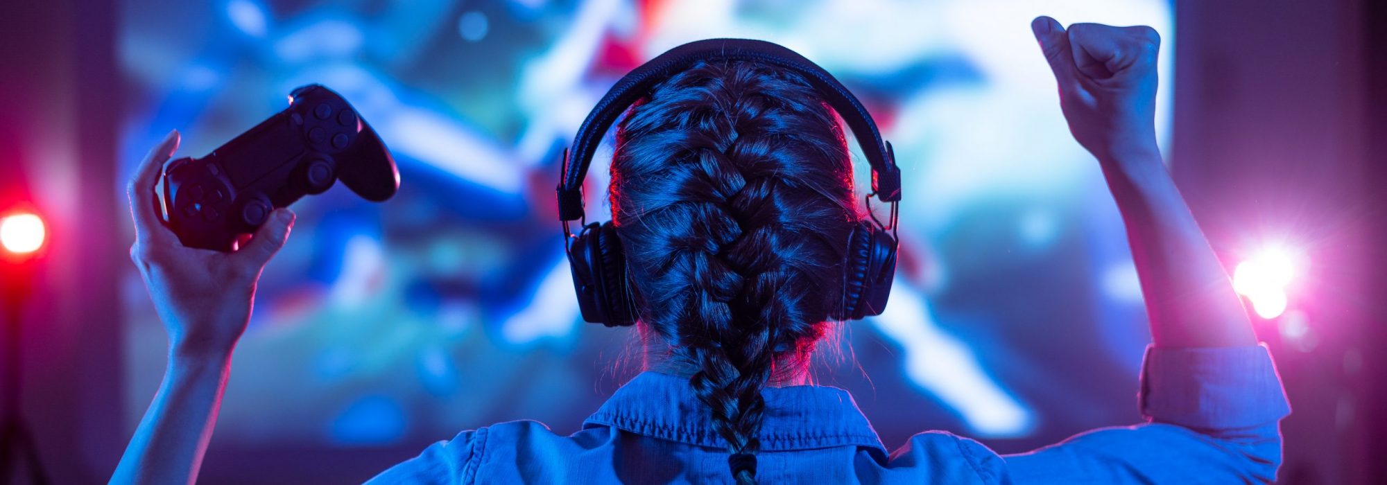 Girl in headphones plays a video game on the big TV screen. Gamer with a joystick. Online gaming with friends, win, prize. Fun entertainment. Teens play adventure games. Back view. Neon lighting