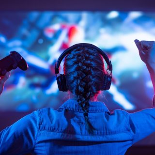 Girl in headphones plays a video game on the big TV screen. Gamer with a joystick. Online gaming with friends, win, prize. Fun entertainment. Teens play adventure games. Back view. Neon lighting